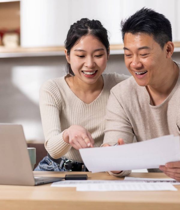 Cheerful asian spouses paying bills on Internet from home, sitting together at kitchen table, holding letters and papers, using computer and smiling, enjoying easy e-banking, copy space